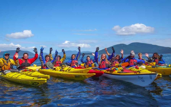 a group of gap year students sit in kayaks and raise their arms in celebration on an outward bound expedition 